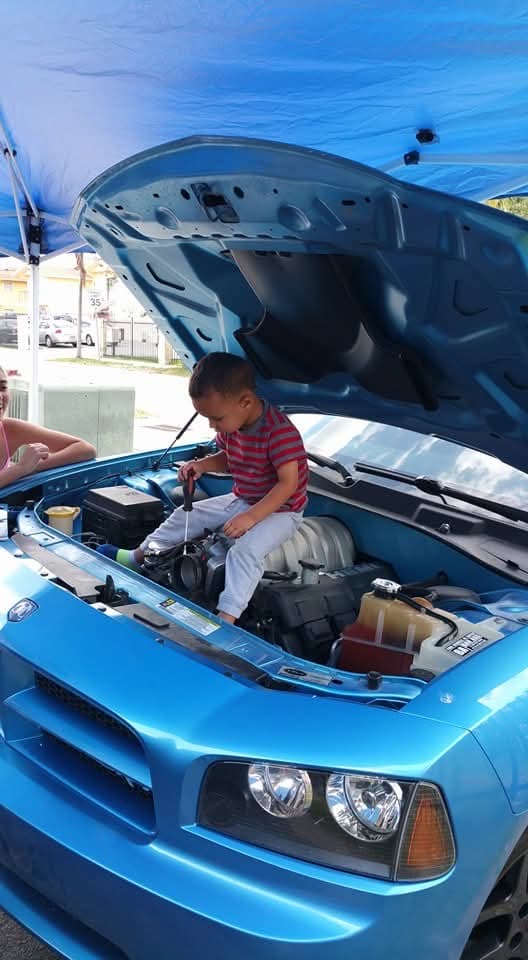 Un niño sentado en el compartimiento del motor de un Dodge Charger azul, con un destornillador en la mano